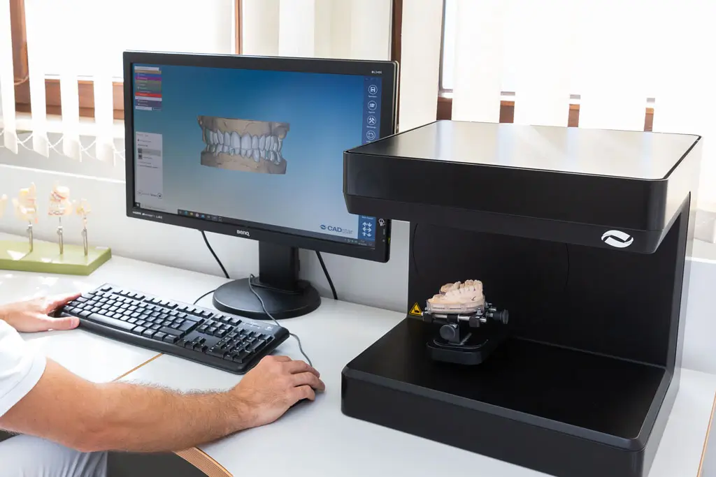 CAD Zahnersatz Mann in weißer Arbeitskleidung sitzt an Schreibtisch und betrachtet 3D-Darstellung von Zähnen auf Computerbildschirm neben Scanner mit Zahnmodell