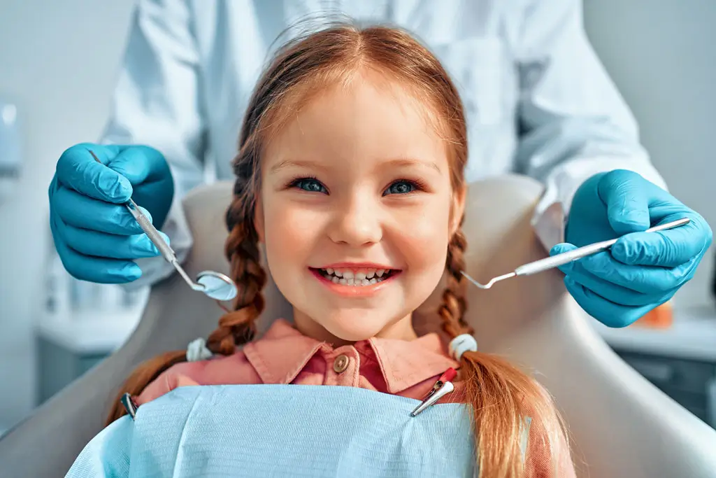 Kind beim Zahnarzt Lächelndes Mädchen mit Zöpfen sitzt beim Zahnarzt, der mit blauen Handschuhen Zahnspiegel und Sonde hält