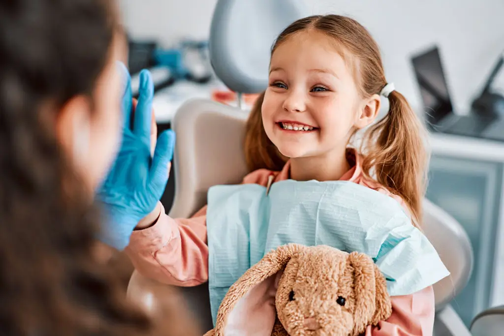 Kinderzahnbehandlung Kind mit Zöpfen und Zahnarztkittel sitzt lächelnd auf Zahnarztstuhl und hält braunes Kuscheltier