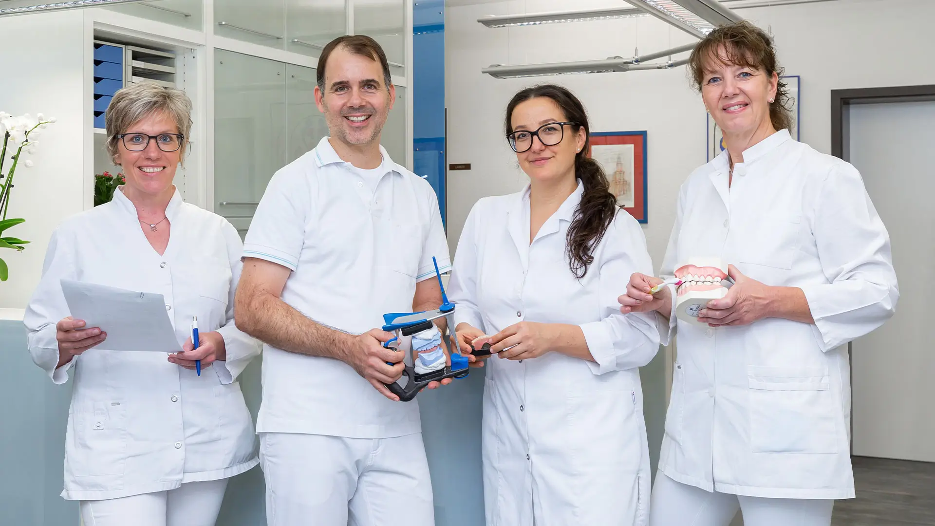 Team Zahnarztraxis Dr. Reimer Drei Zahnärztinnen und ein Zahnarzt in weißer Arbeitskleidung stehen in einer Praxis und halten verschiedene zahnmedizinische Modelle und Instrumente