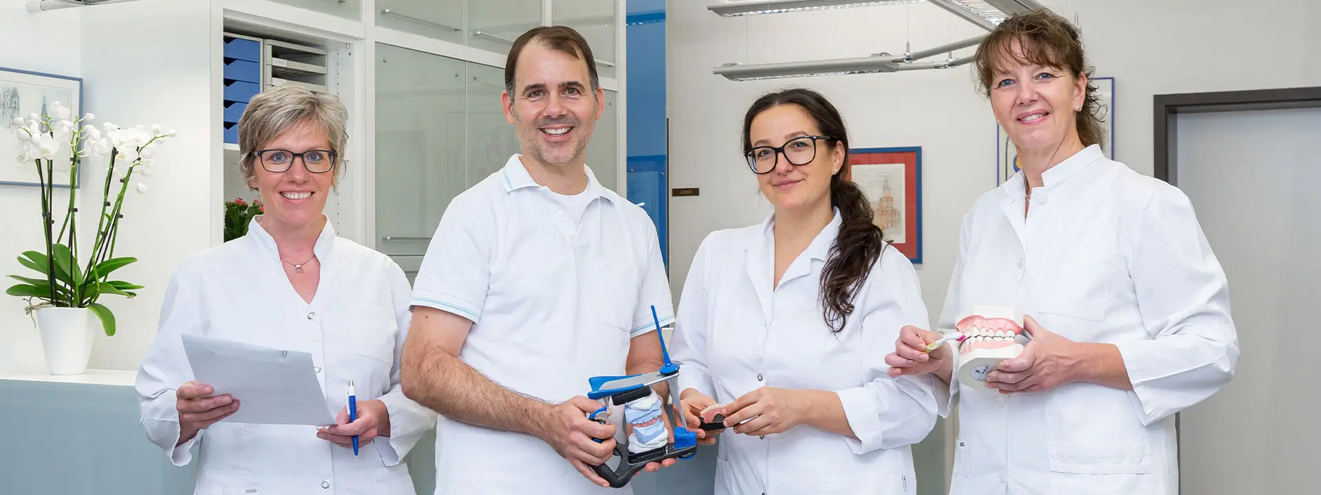 Team Zahnarztraxis Dr. Reimer Drei Zahnärztinnen und ein Zahnarzt in weißer Arbeitskleidung stehen in einer Praxis und halten verschiedene zahnmedizinische Modelle und Instrumente