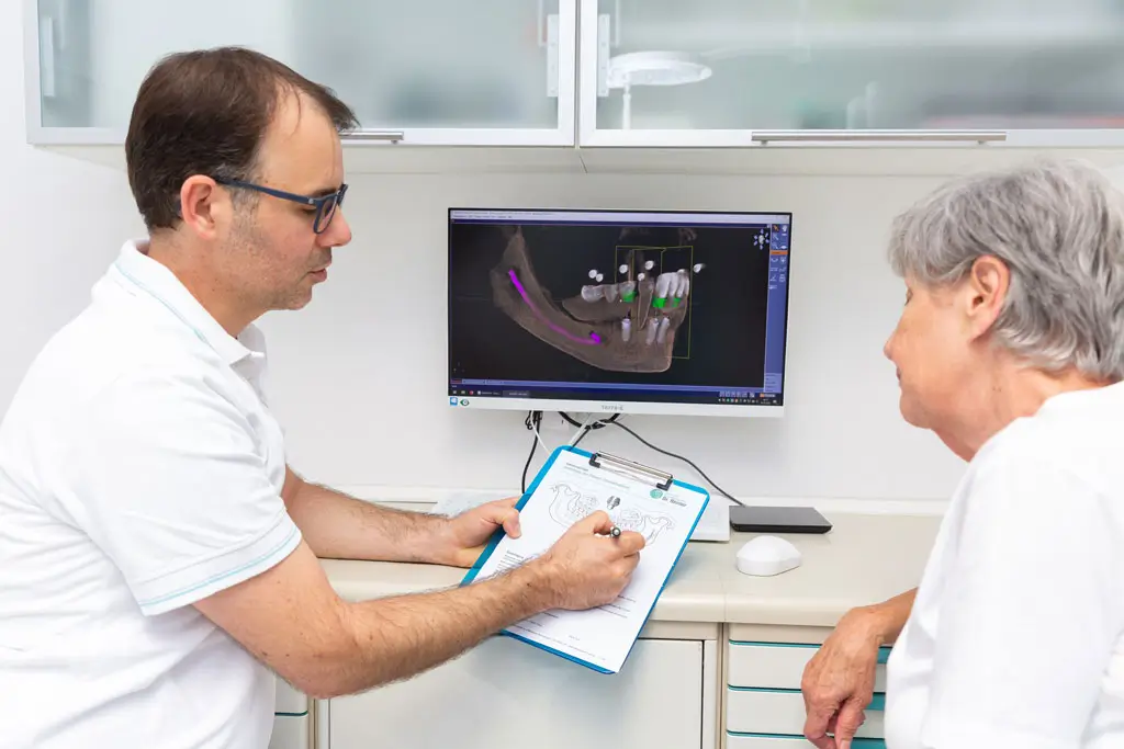Zahnersatz Beratung Zahnarzt erklärt älterer Patientin anhand eines Kieferröntgenbilds auf einem Monitor und einem Klemmbrett mit Zeichnungen