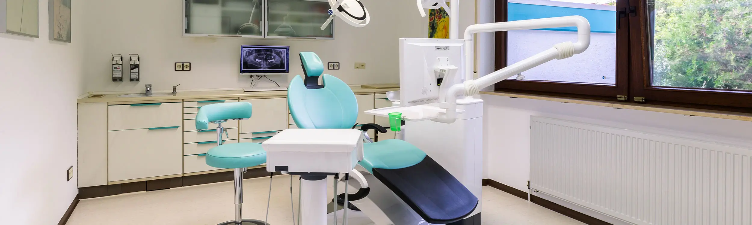 Zahnarztstuhl im Behandlungszimmer Zahnarztstuhl mit blauem Sitz und Rückenlehne in einem hellen Behandlungsraum mit Fenster und weißen Schränken